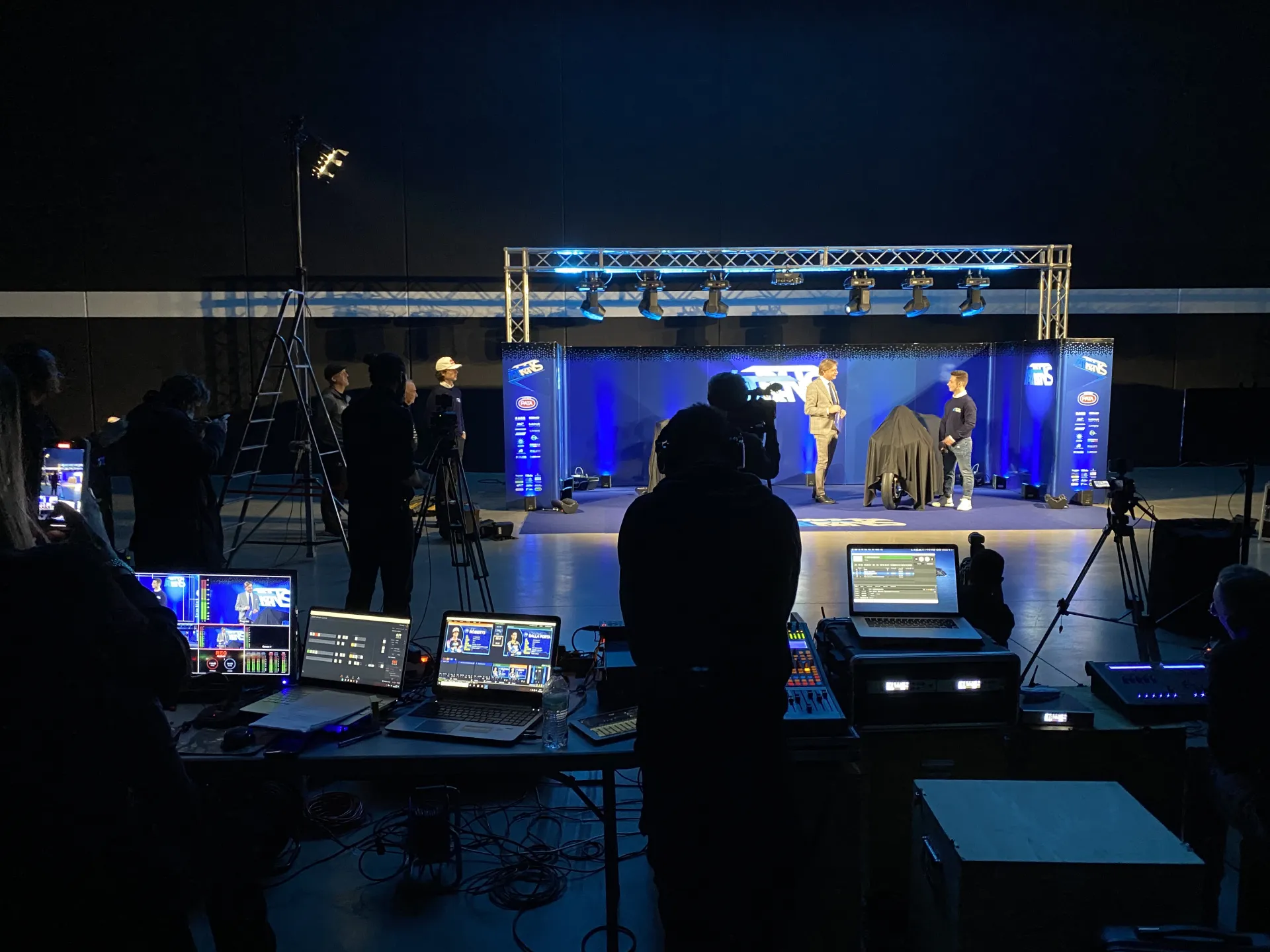 Postazione regia con schermi, laptop e palco illuminato durante un evento corporate (Victor Music)